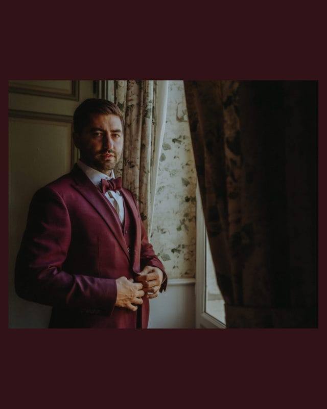 Geoffrey ajuste sa veste bordeaux près d’une fenêtre au château de la Berchère, pendant les préparatifs de son mariage à Nuits-Saint-Georges.