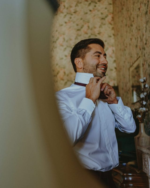 Geoffrey sourit en ajustant son nœud papillon bordeaux devant un miroir, pendant les préparatifs au château de la Berchère.